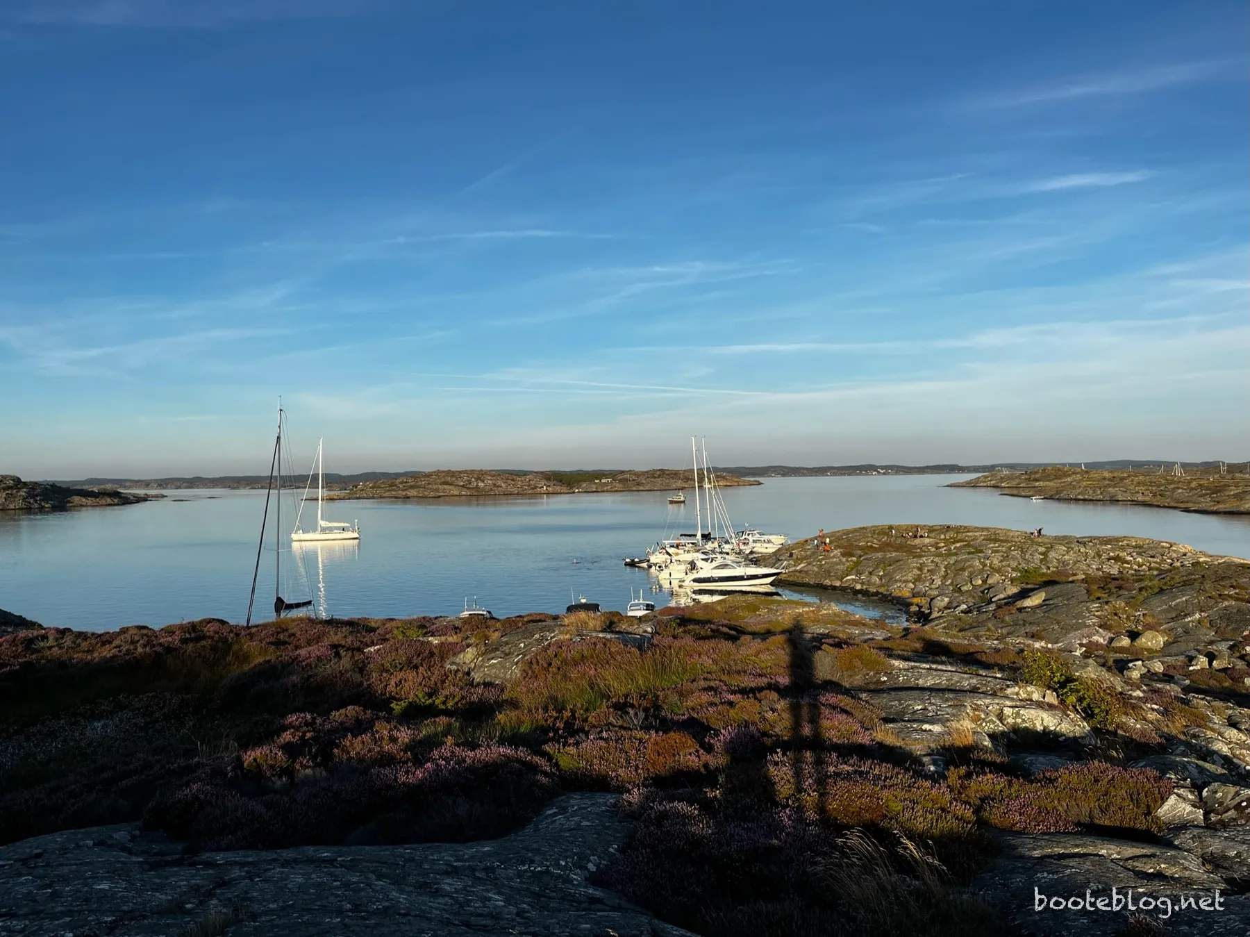 Blick von einer felsigen Anhöhe auf eine geschützte Bucht in den Stockholmer Schären: Segelboote und Yachten liegen zwischen Granitfelsen mit Heidekraut, im Hintergrund weitere Inseln.