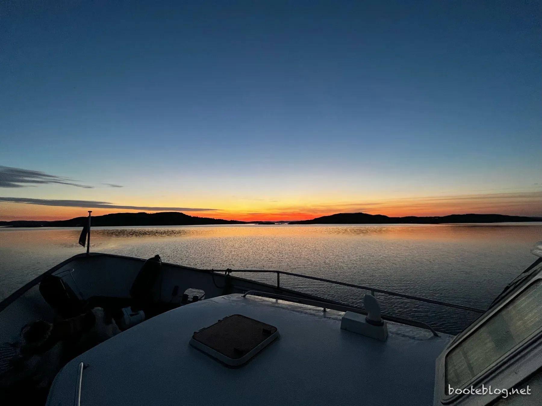 Blick vom Vorschiff eines Boots auf einen Sonnenuntergang über dem ruhigen Wasser der schwedischen Schären, Inseln als Silhouetten am Horizont, der Himmel von Orange zu Blau.
